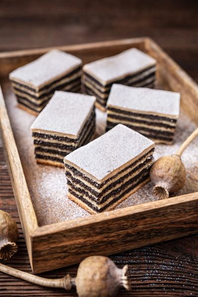 Squares of a layered cake with dark poppy seed fillings, dusted with powdered sugar, in a wooden tray